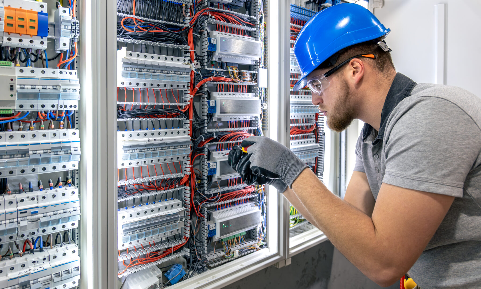 An electrical technician installs components inside a switchboard panel. Artificial lighting, one person, indoor setting, precise technical work.